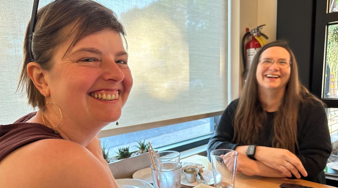 Two women smiling and laughing while sitting at a table near a window in a casual dining setting.