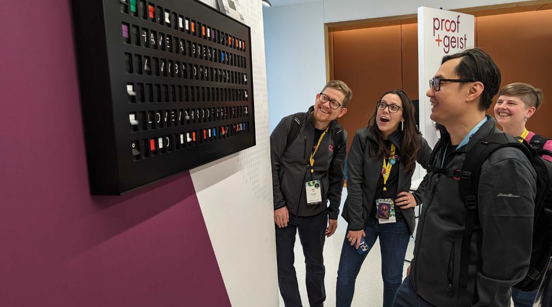 Four people wearing conference badges look surprised and amused while viewing a black display board with colored squares and text on a white wall.