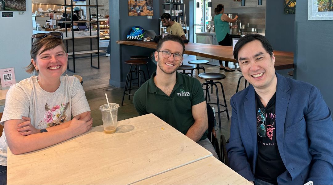 Three smiling adults sitting at a light wooden table in a cafe with a coffee shop counter in the background.