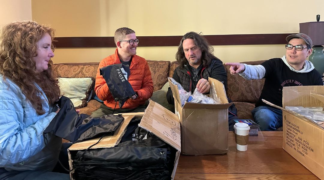 Four people sitting on a couch surrounded by open cardboard boxes, unpacking black bags labeled 'PAUSE.'