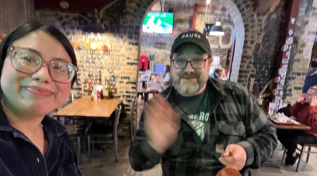 A woman with glasses smiling at the camera and a man wearing glasses and a 'PAUSE' cap waving inside a rustic restaurant.
